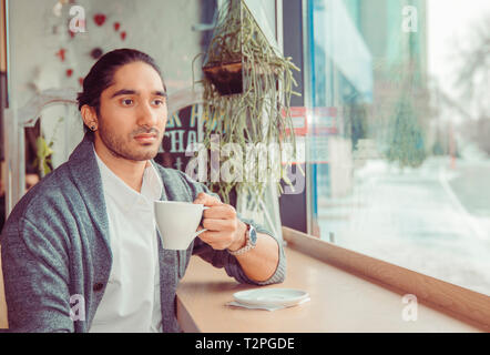 Triste riflessivo uomo che guarda in basso pensieroso coppa di ritegno. Closeup ritratto di un bel ragazzo indossa abbigliamento casual seduta vicino alla finestra Tabella di caffè Foto Stock