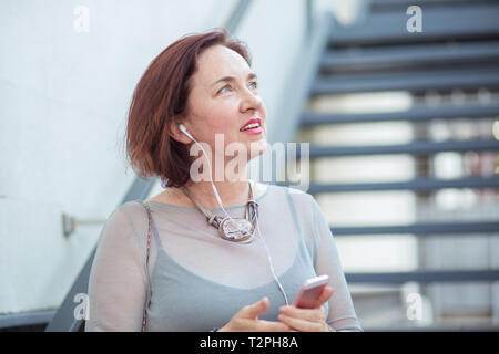 Felice donna matura fantasticando parlando al telefono per ascoltare musica in cuffia isolato paesaggio urbano all'aperto scale sullo sfondo Foto Stock