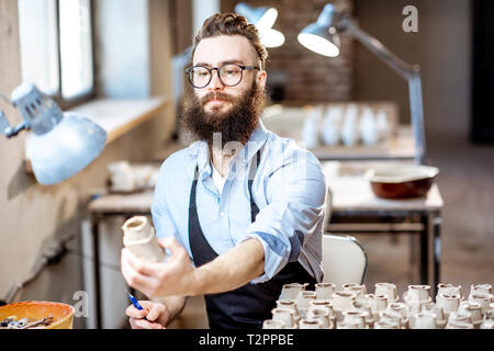 Bel uomo barbuto lavora con pezzi di ceramica al posto di lavoro del negozio di ceramiche Foto Stock