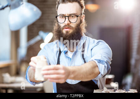 Bel uomo barbuto lavora con pezzi di ceramica al posto di lavoro del negozio di ceramiche Foto Stock