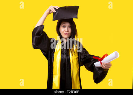 Graduate con un diploma in mano, smorfie e si rallegra per il successo del giorno di graduazione, Immagine su sfondo giallo Foto Stock