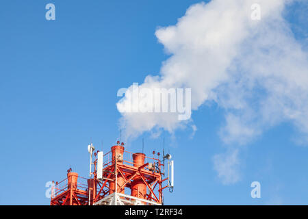White nocivi del fumo proveniente al di fuori del rosso-e-bianco con tubi di comunicazione mobile antenne in una fabbrica nel centro della città contro lo sfondo di Foto Stock