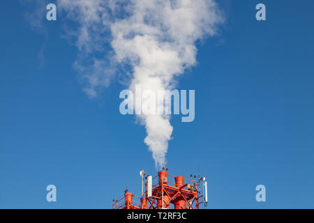White nocivi del fumo proveniente al di fuori del rosso-e-bianco con tubi di comunicazione mobile antenne in una fabbrica nel centro della città contro lo sfondo di Foto Stock