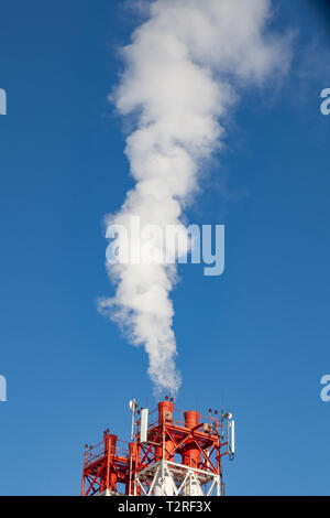 White nocivi del fumo proveniente al di fuori del rosso-e-bianco con tubi di comunicazione mobile antenne in una fabbrica nel centro della città contro lo sfondo di Foto Stock