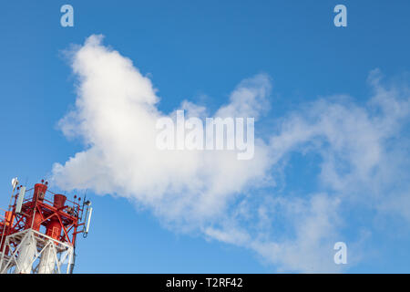 White nocivi del fumo proveniente al di fuori del rosso-e-bianco con tubi di comunicazione mobile antenne in una fabbrica nel centro della città contro lo sfondo di Foto Stock