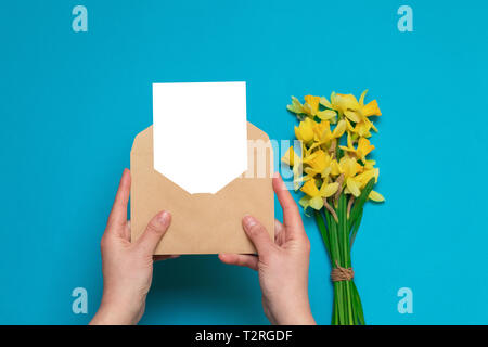 Mani craft busta con una scheda, freschi daffodils giallo su sfondo blu. Scrivania da ufficio, concetto di primavera. Foto Stock
