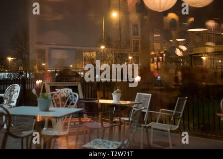 Vista interna del bar/bistrot/caffè alla moda, in serata, dall'esterno della strada. Sedie e tavoli esterni visibili nella finestra di riflessione. Foto Stock