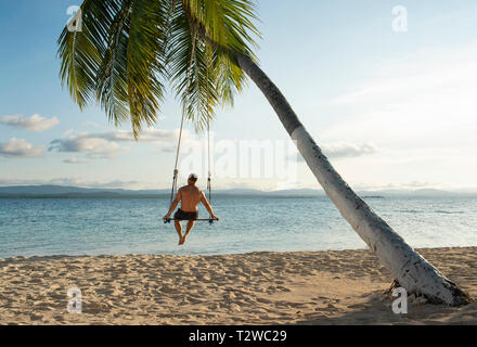 Vista posteriore dell'uomo in una spiaggia swing attaccato a un albero di palma in isole San Blas. Destinazione di viaggio, stile di vita / Concetto di vacanza. Panama, Ott 2018 Foto Stock