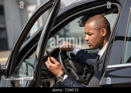 I giovani africani imprenditore americano seduto in macchina porta aperta trattenendo il volante con una mano il controllo navigazione sul serio dello smartphone Foto Stock