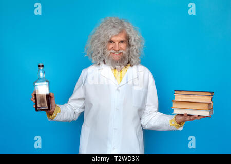 Vecchio con i capelli lunghi uomo che indossa Doctor's vestaglia studio isolato sulla parete blu in piedi tenendo la bottiglia di alcool in una mano e la pila di libri in un altro gabinetto Foto Stock