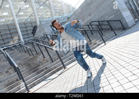 Giovane americano africano uomo che indossa le cuffie per ascoltare musica ballare vicino a scale sulla strada di città azienda smartphone ridendo allegro Foto Stock