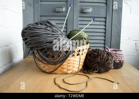 Traditional colorful yarns for hobby knitting in a straw eco basket on a light wooden surface, knitting needles, knitted basket. Foto Stock