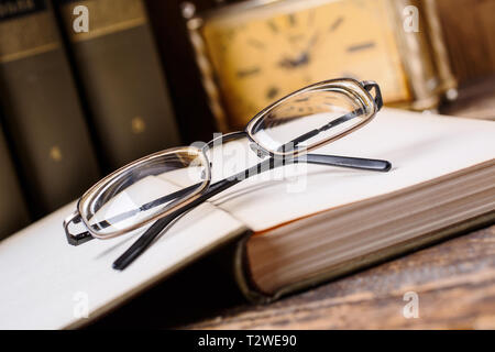 Dopo la lettura, smontare gli occhiali e lasciate che i vostri occhi di riposo. I vetri si trovano sul libro aperto sullo sfondo di libri e il vecchio orologio. messa a fuoco selettiva Foto Stock