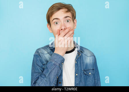 Funny giovane adolescente di sesso maschile in denim shirt su sfondo blu in orrore copre la bocca Foto Stock