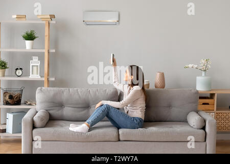 Giovane donna sorridente seduto sul divano di accendere il condizionatore d'aria Foto Stock