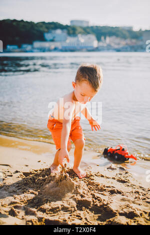 Oggetto di costruzione e di industria pesante. Astrazione bambino ragazzo giocando sulla sabbia vicino al fiume in estate toy rosso modello trattore, escavatore machi Foto Stock