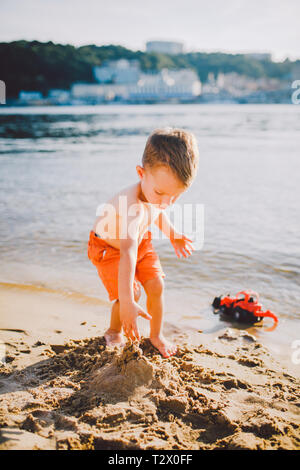 Oggetto di costruzione e di industria pesante. Astrazione bambino ragazzo giocando sulla sabbia vicino al fiume in estate toy rosso modello trattore, escavatore machi Foto Stock