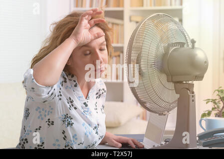 La donna soffre di calore mentre si lavora in ufficio e si tenta di raffreddarsi dalla ventola Foto Stock