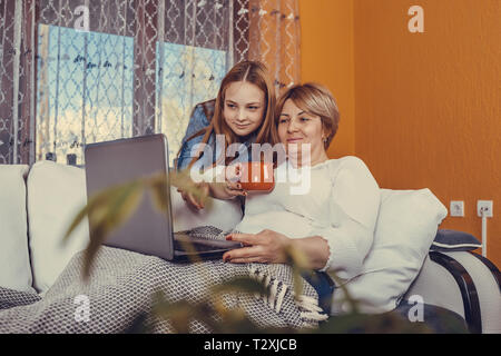 Sorridente madre e figlia seduti sul divano guardando un video su laptop, teen girl appendere fuori con la sua madre, rilassatevi sul divano usando il computer, mamma e bambino resto Foto Stock