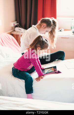 Ragazza adolescente tramite telefono cellulare insieme con la sua sorellina la visione di film di animazione sui tablet, entrambe ragazze seduta nel letto in camera da letto Foto Stock