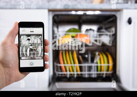 Close-up di mano di una persona operante lavastoviglie con lo smartphone Foto Stock