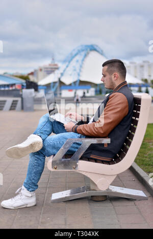 Giovane ragazzo in giacca di pelle e i jeans. freelancer lavora al computer su strada. l uomo si siede sul banco. profilo vista laterale Foto Stock