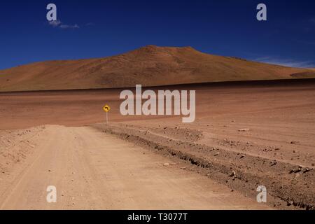 Strada sterrata attraverso il red wasteland contrastante con il profondo blu del cielo senza nuvole, isolato perso il cartello giallo che mostra direzione sinistra - deserto di Atacama, Cile Foto Stock