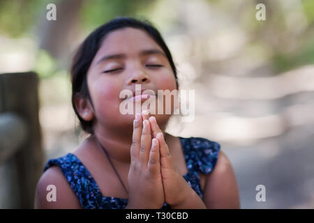 Nativo di una ragazza giovane con le mani insieme nella preghiera, in un ambiente esterno alla preghiera a Dio con un sottile sorriso. Foto Stock