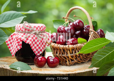 Un vasetto di marmellata e cesto di ciliegie dolci su sfondo nel giardino all'aperto. Foto Stock