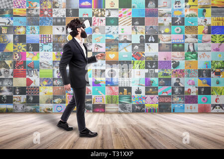 Uomo Barbuto con gli occhiali di realtà virtuale, passeggiate in una stanza vuota, un collage di foto a colori e video sulla parete, concetto di navigazione Foto Stock