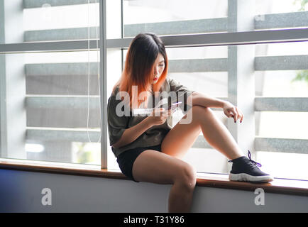 Asian highschool ragazza seduta dalla finestra utilizza lo smartphone Foto Stock