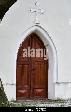 Porta della chiesa in legno adagiata in un arco gotico su una facciata bianca, con sopra una croce cristiana e un tronco d'albero in primo piano. Foto Stock