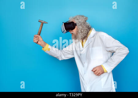 Vecchio con i capelli lunghi uomo che indossa Doctor's vestaglia e realtà virtuale cuffie studio isolato su blu a piedi della parete di contenimento agressive martello vista laterale Foto Stock