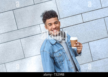 Giovane mulatto uomo in piedi indossando le cuffie attorno al collo isolati su parete in cemento azienda tazza di caffè caldo in cerca della telecamera gioioso sorridente Foto Stock