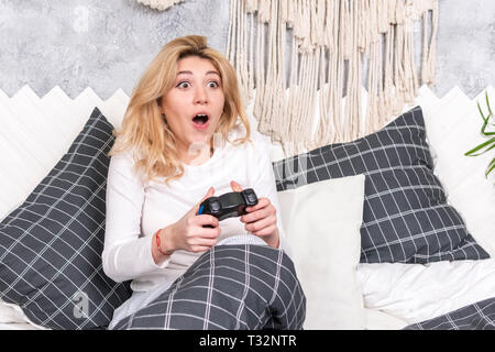 Bella donna di gioco d'azzardo gioca il gioco con il joystick seduta sul letto Foto Stock