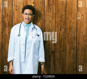 Giovane africano medico americano donna che indossa medical rivestire sfondo isolato in shock viso, cercando scettici e sarcastico, sorpreso con open Foto Stock