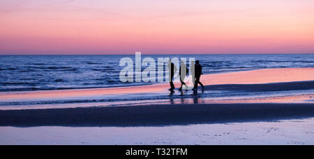 Quattro irriconoscibile giovani uomini camminando lungo il mare su una deserta spiaggia sabbiosa, essi sono vicini tra loro parlando non vi è una rosa glow in the sky riflettendo Foto Stock