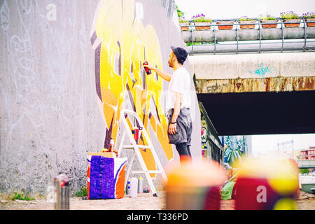 Artista di strada di verniciatura di un coloratissimo graffito su un muro grigio sotto il ponte - Giovani graffiter scrittura e disegno murales con spray di colore giallo Foto Stock