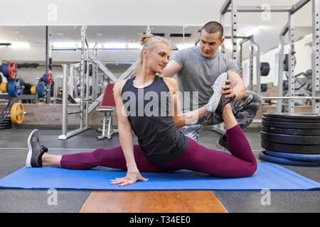 Giovane forte uomo muscolare aiutando i giovani donna atletica stretching sul materassino yoga in palestra. Foto Stock