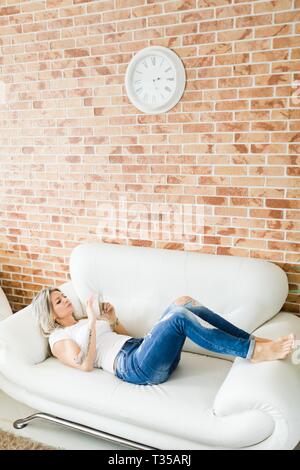 Giovane donna in jeans utilizzando smart phone comodamente sdraiati sul divano bianco. Muro di mattoni in background. Foto Stock
