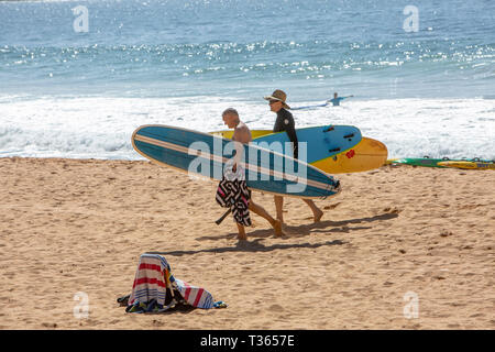 Due uomini camminare sulla sabbia a Palm Beach che trasportano le loro tavole da surf longboard, Sydney, Australia