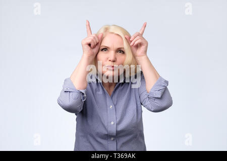Divertenti scherzi senior donna bionda rendendo le corna con le dita. Studio shot Foto Stock