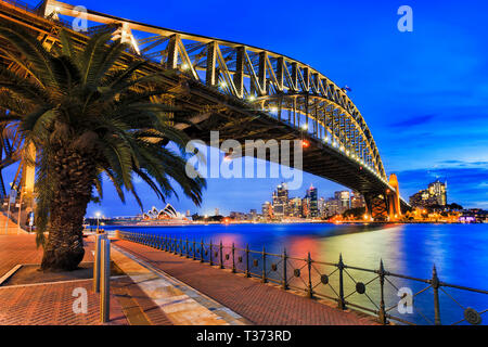 Vista laterale di acciaio arcata del ponte del Porto di Sydney durante il blu ora a Sunset over sfocato acque del porto di Sydney verso le rocce e il CBD di Città w Foto Stock