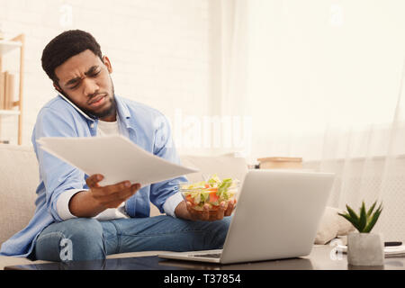 Non è tempo di pausa. Sistema nervoso african american guy mangiare sano colazione, parlando al telefono cellulare e di lavoro sul portatile a casa, spazio di copia Foto Stock