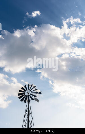 Basso angolo vista di una vecchia, multi-bladed, metallo pompa di vento in controluce contro il cielo blu con nuvole bianche. Foto Stock