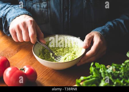 L'uomo schiacciare l'avocado nel recipiente, tavolo in legno sfondo. Preparazione alimentare immagine, sano e vegano cibo vegetariano Foto Stock