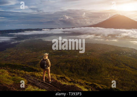 L'uomo guarda turistica all'alba sul vulcano Batur sull isola di Blai in Indonesia. Escursionista uomo con zaino travel sulla sommità del vulcano, viaggi Foto Stock