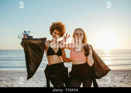 Due femmina surfers dopo il prelievo di lettiera sulla spiaggia. Volontari di sesso femminile dopo la pulizia della zona spiaggia, in piedi insieme sorridente e gesticolando vict Foto Stock