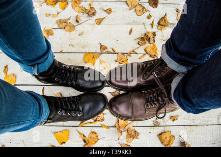 Elegante giovane. Vista superiore del paio di piedi in eleganti stivali in piedi insieme sul molo in legno con foglie di giallo. Coppia giovane in piedi vicino a ciascuno altri Foto Stock
