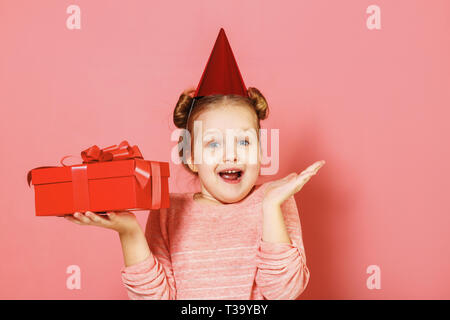 Closeup ritratto di una bambina con ciuffi di capelli su sfondo rosa. Il bambino ha un cappello sulla sua testa e contiene una scatola con un dono nelle sue mani. Foto Stock